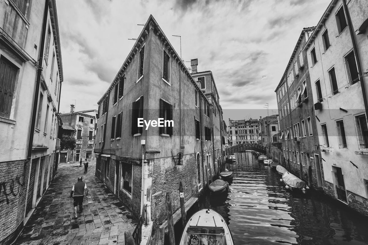 Boats moored on canal amidst building against cloudy sky