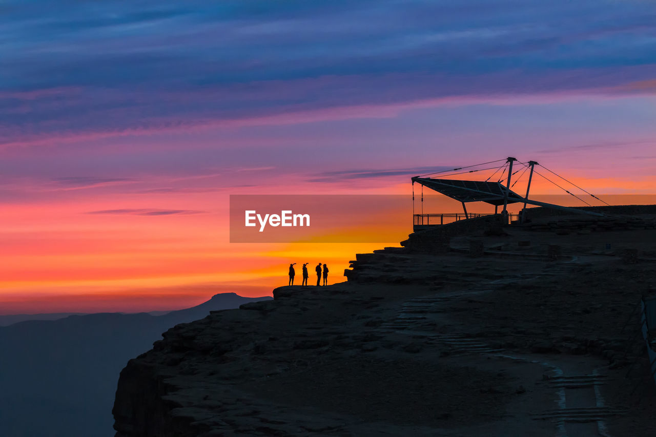 Silhouette people standing on cliff against orange sky