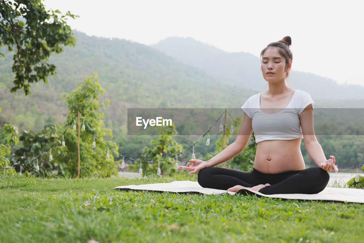 Full length of pregnant young woman meditating while sitting on mat over grass