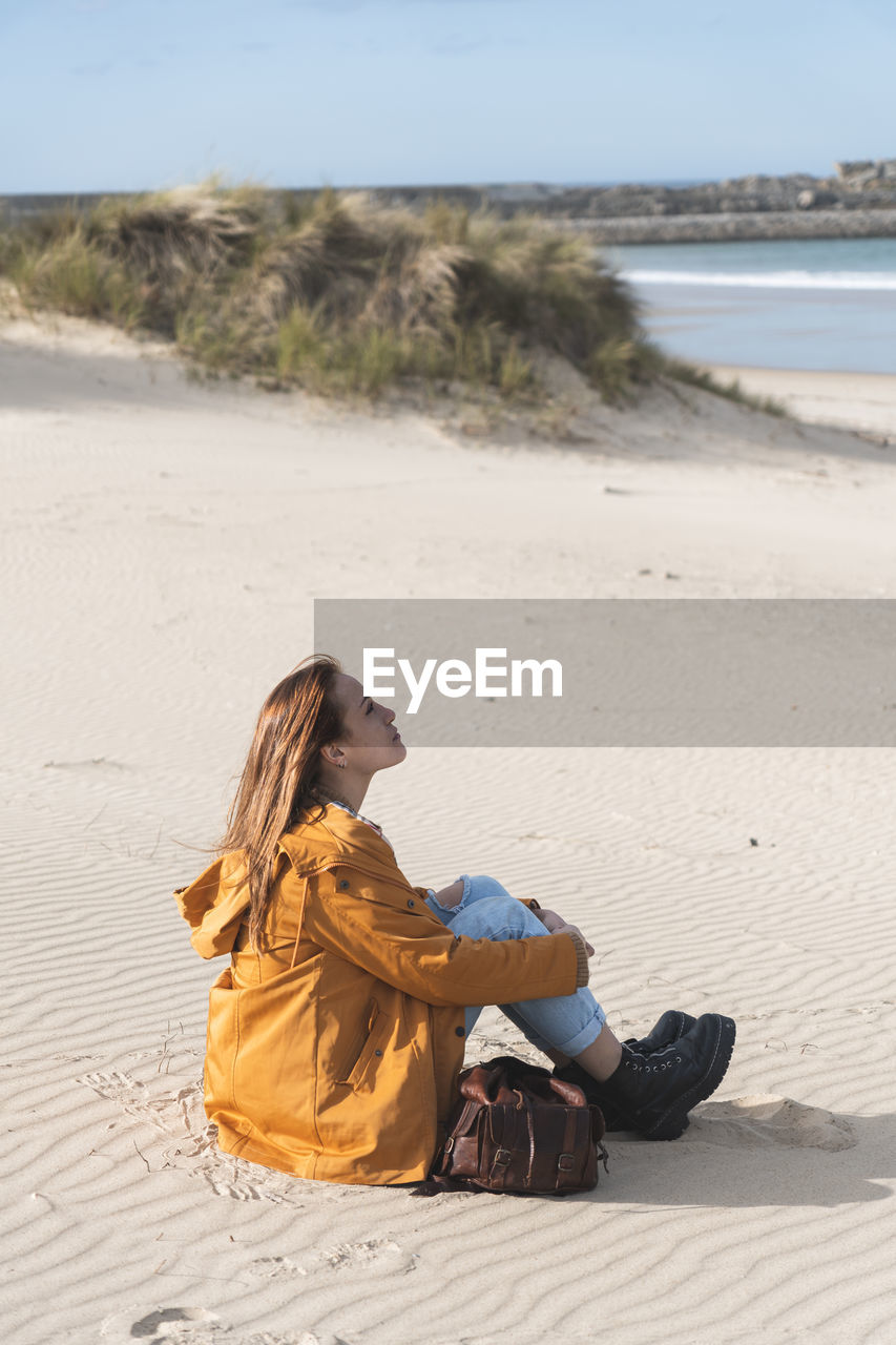 Young woman hugging knees while sitting at beach during sunny day