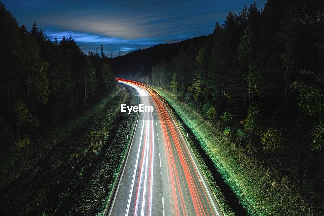 Light trails on road amidst trees against sky at night