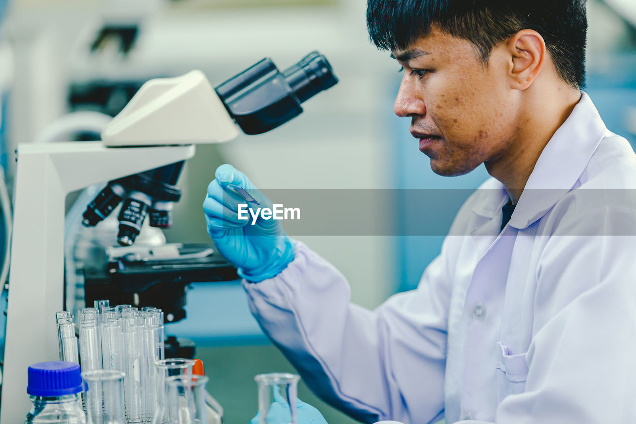 Asian man lab technician in protective glasses and gloves sits next to a microscope in laboratory.