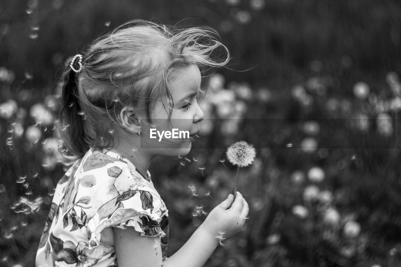 Side view of girl standing holding dandelion against blurred background