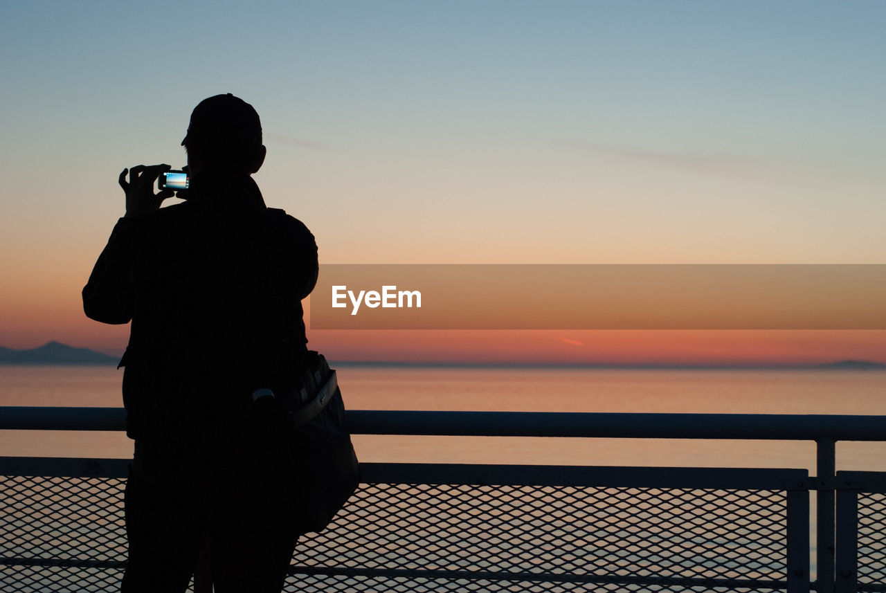 Silhouette man photographing sea against sky during sunset