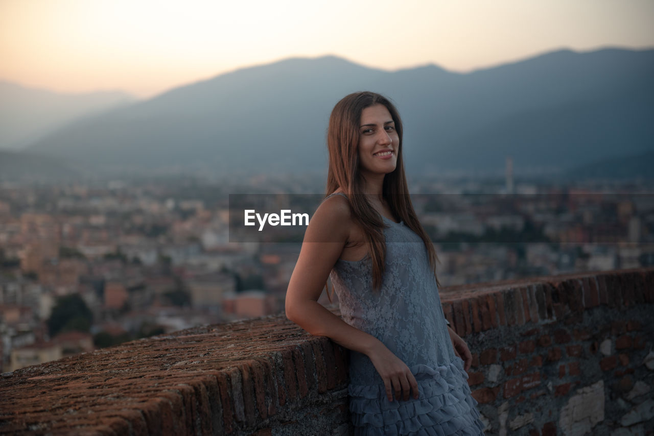 Portrait of young woman in city against sky during sunset