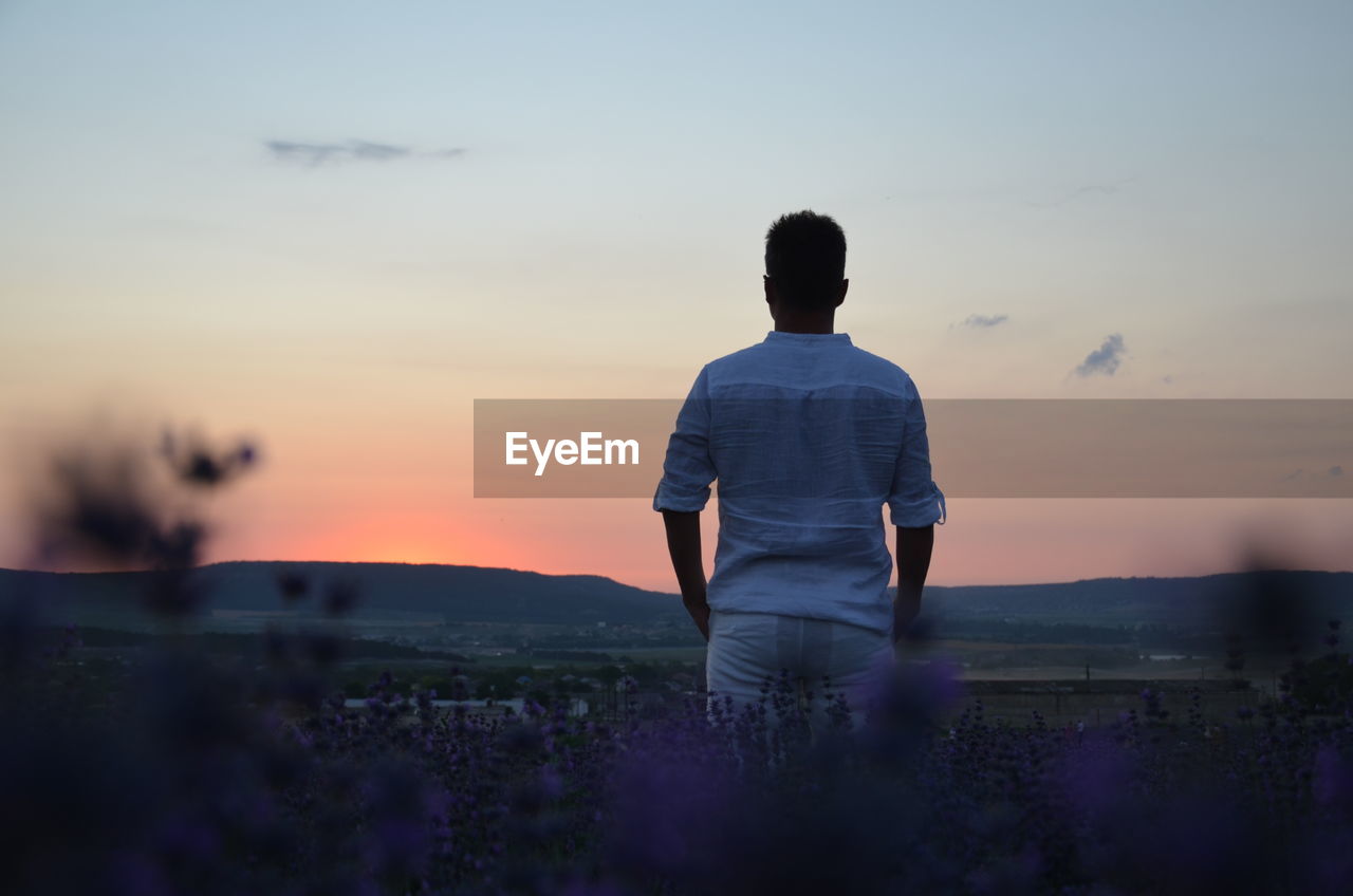 Rear view of man standing on field against sky during sunset