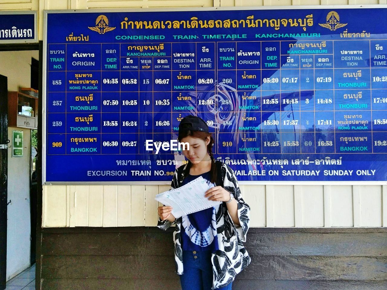 Woman reading document while standing against arrival departure board at railroad station