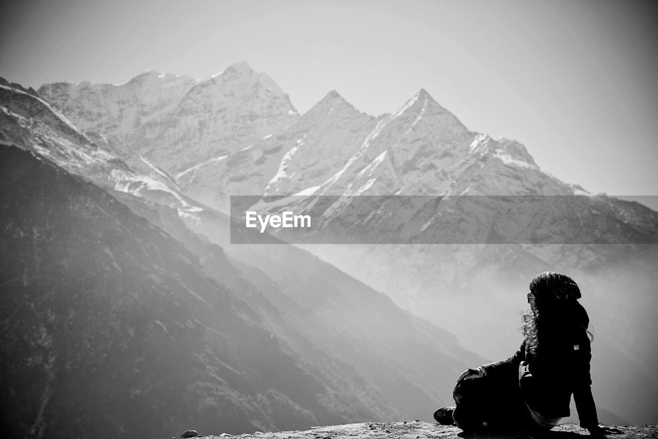 Woman sitting on rock by snowcapped mountains in foggy weather
