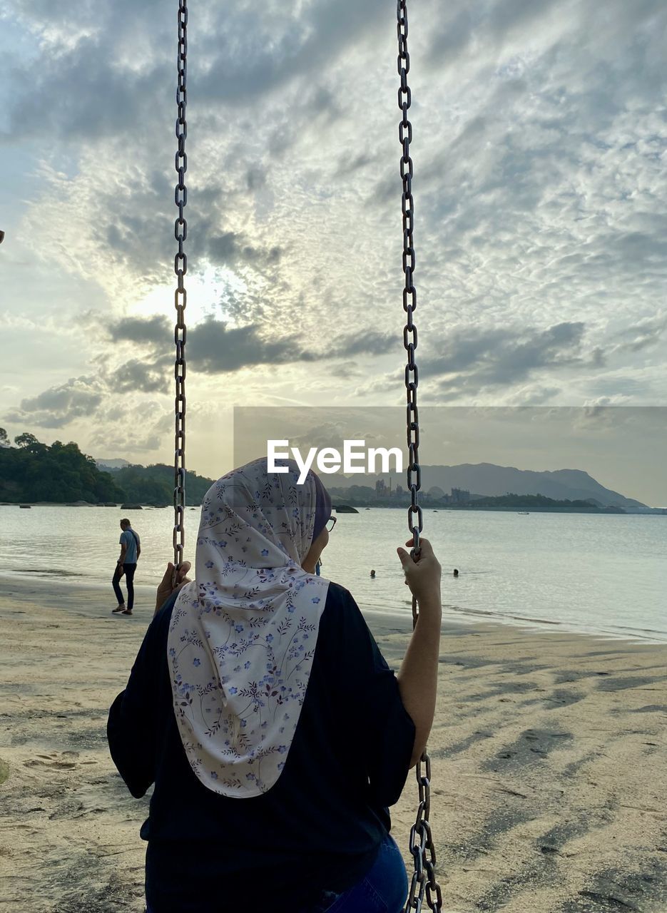sky, swing, sea, land, beach, water, nature, cloud, chain, one person, sand, adult, lifestyles, scenics - nature, day, blue, leisure activity, outdoors, beauty in nature, women, ocean, rear view, rope, sitting, sunlight, men, childhood, playground, child, person, tranquility, tranquil scene, hanging