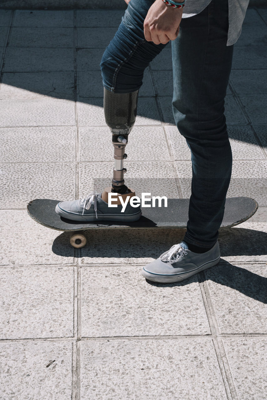 Side view of young guy with leg prosthesis standing and riding skateboard on city street