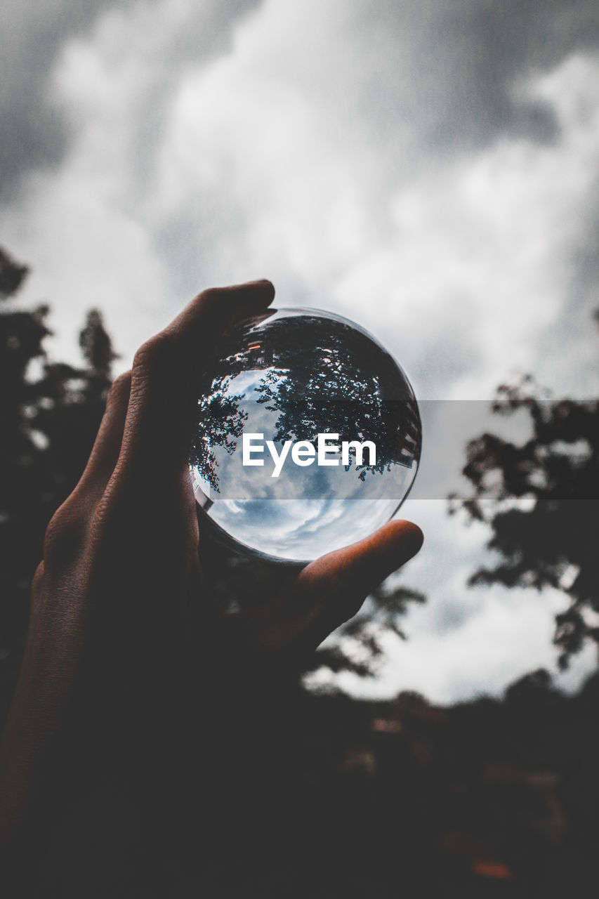 Close-up of hand holding crystal ball against cloudy sky