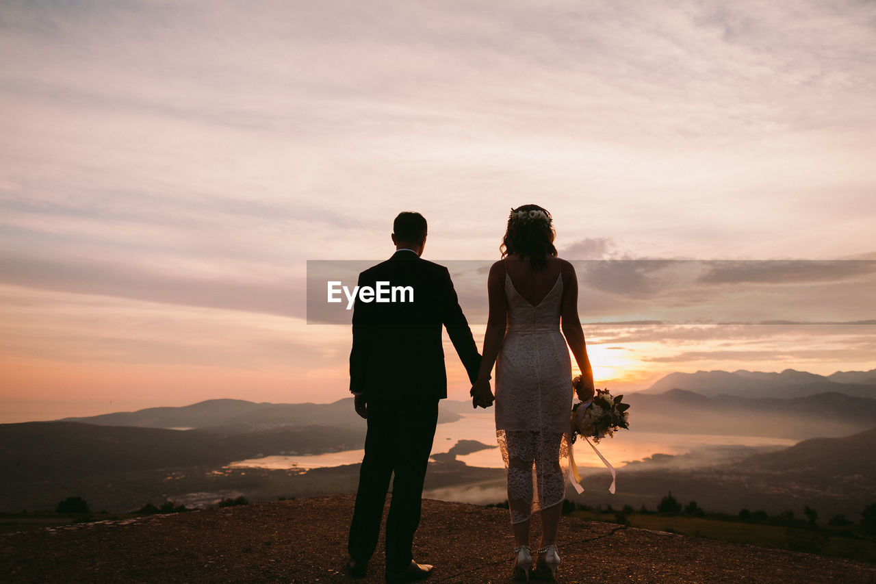 FRIENDS STANDING ON SHORE AGAINST SKY AT SUNSET