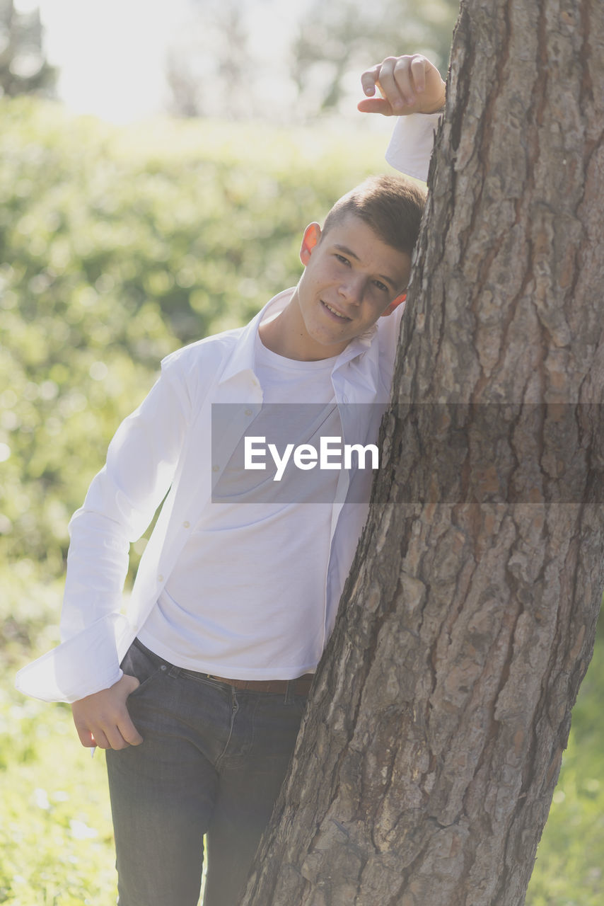 Portrait of teenage boy standing by tree trunk at park