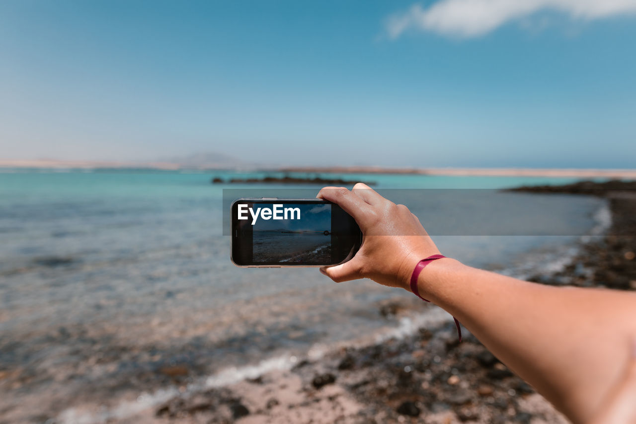 Crop unrecognizable female tourist demonstrating photography of ocean on cellphone under cloudy blue sky during summer trip