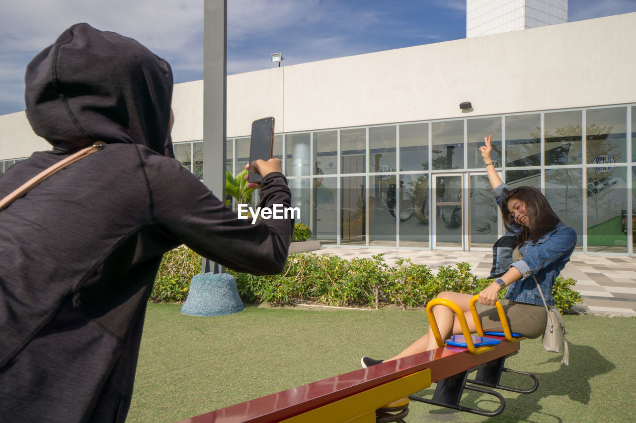 Rear view of man photographing woman sitting on seesaw against built structure