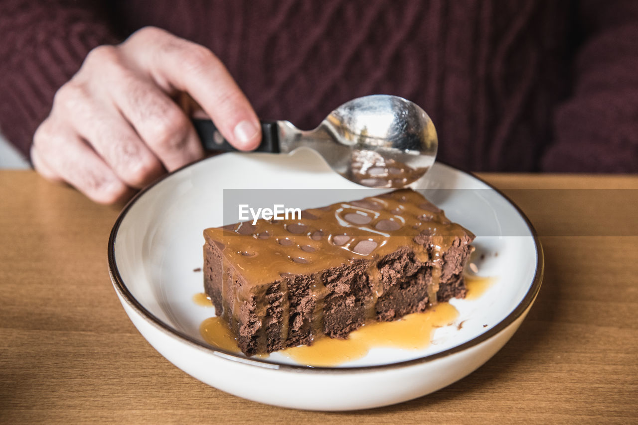 Close-up of hand holding spoon over dessert in plate on table