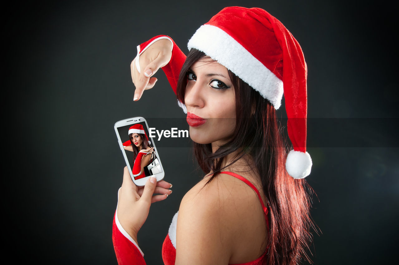 Portrait of smiling woman in santa costume using mobile phone on black background