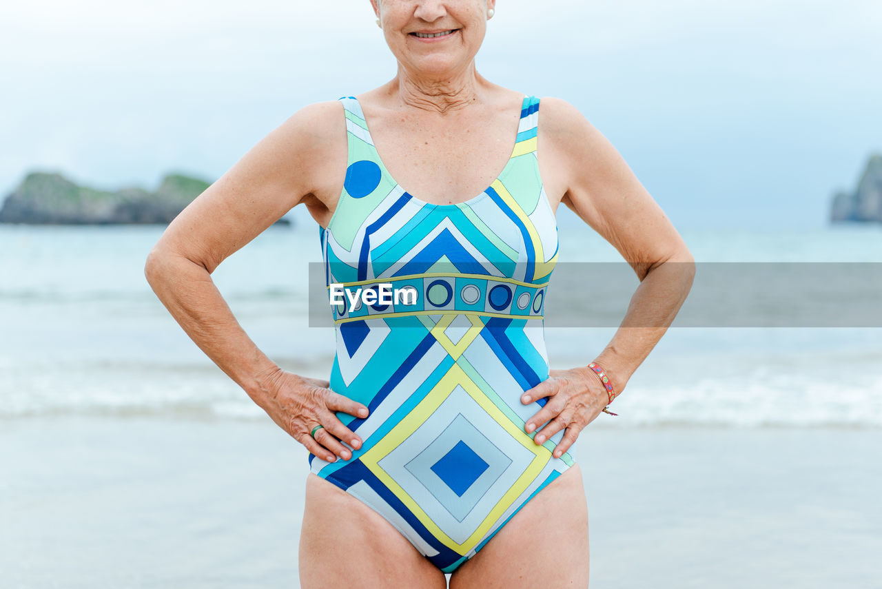 Crop aged female with fit body wearing stylish colorful swimsuit with geometric print standing with hands on waist against sea in summer day