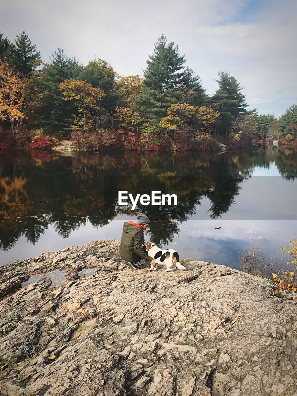 Man crouching with dog on rock by lake against sky