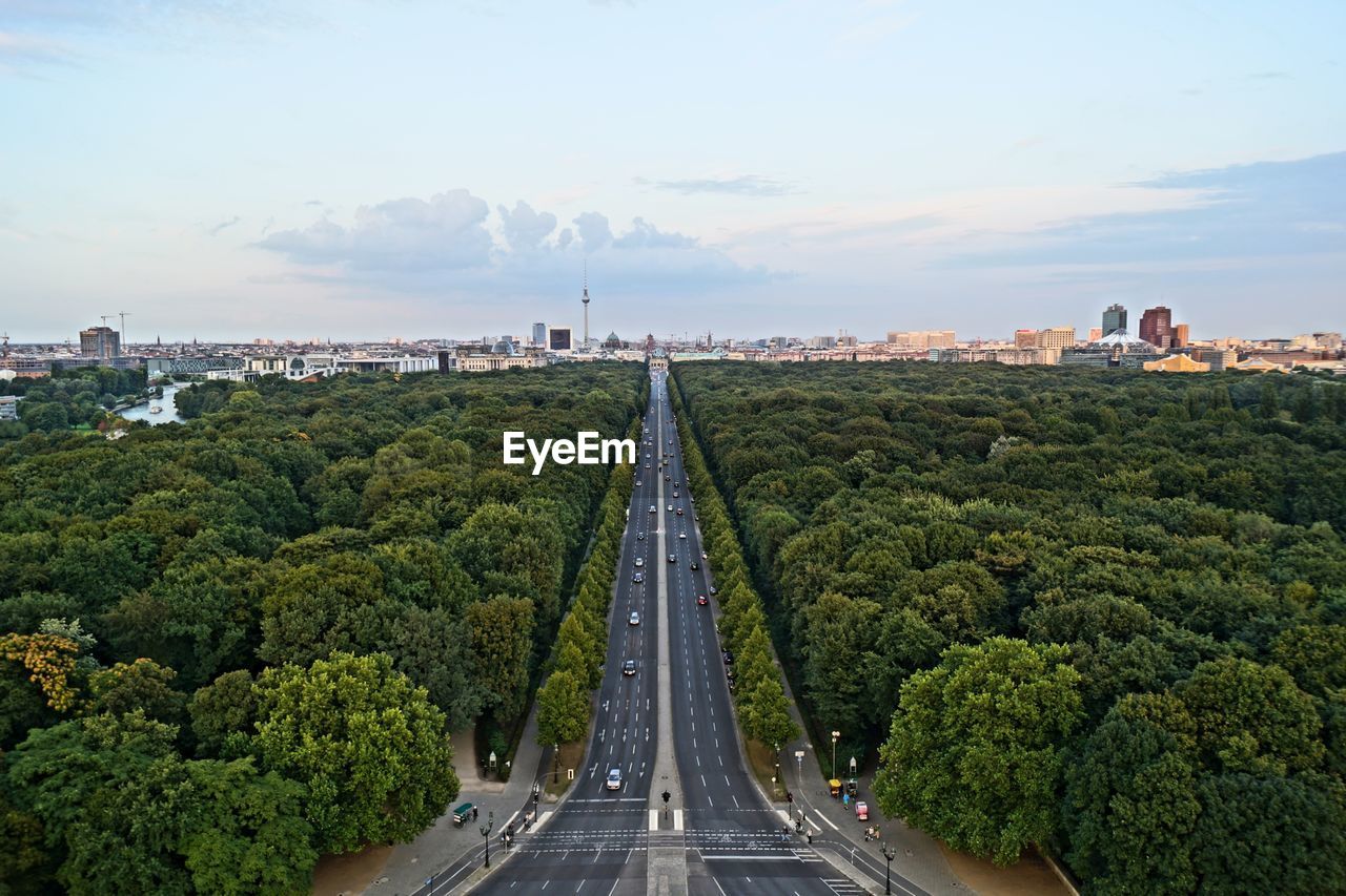 Street amidst trees leading towards city against sky