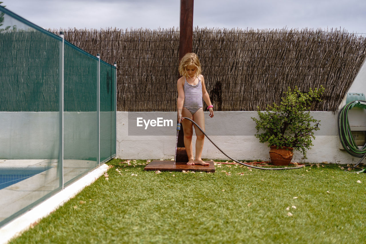 Cute little girl in swimsuit walking and watering green lawn from hose during summer holidays in countryside