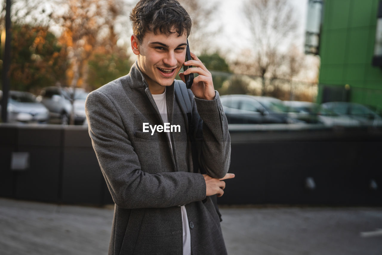 telephone, wireless technology, mobile phone, using phone, communication, technology, one person, adult, men, business, businessman, portable information device, smartphone, city, standing, young adult, architecture, talking, emotion, waist up, street, formal wear, smiling, business finance and industry, blazer, happiness, looking, listening, car, person, portability, transportation, city life, answering, motor vehicle, spring, corporate business, outdoors, cheerful, lifestyles, clothing, holding, occupation, mode of transportation, day, front view, jacket, nature, focus on foreground, vehicle, brown hair