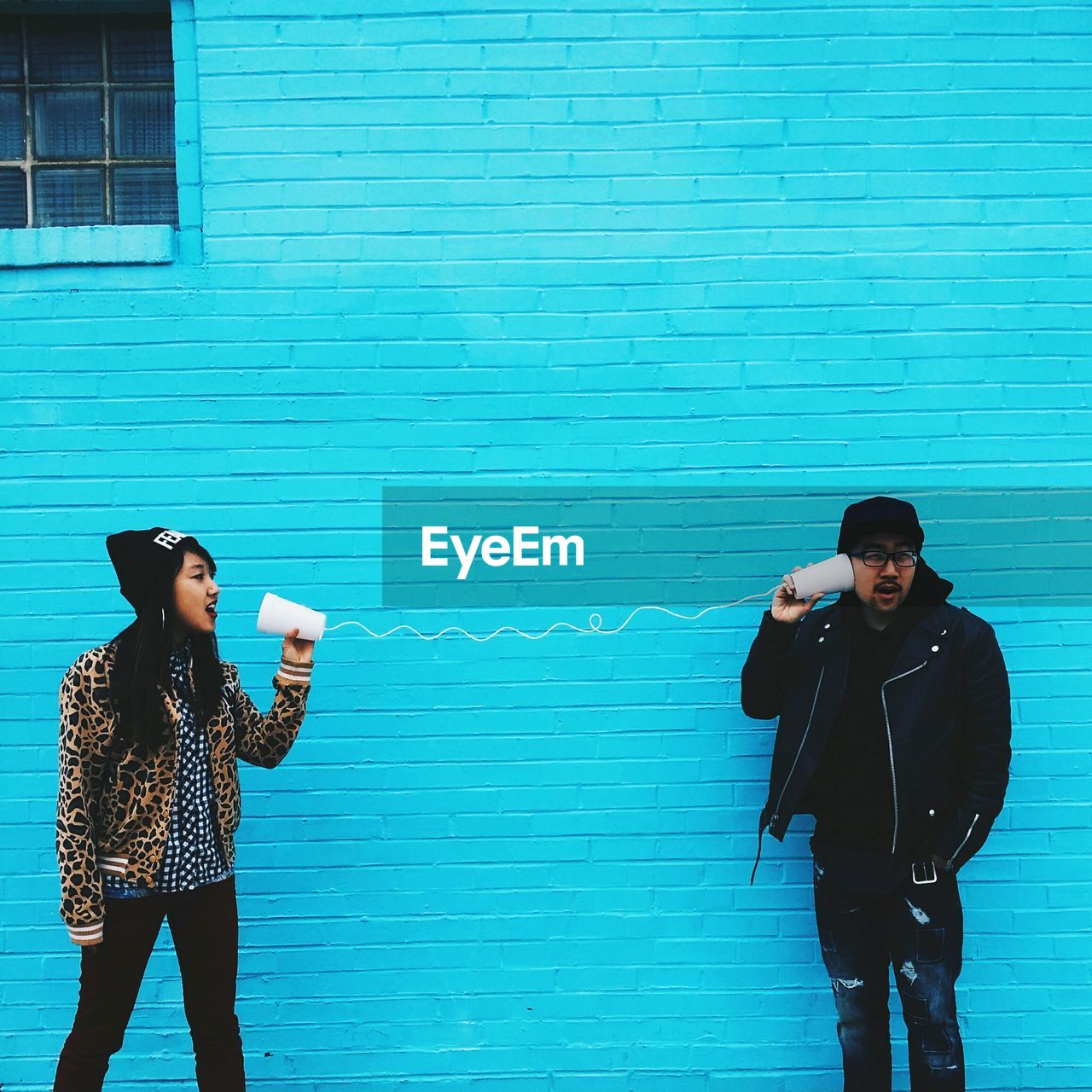 Man and woman using cups and string to communicate against blue wall