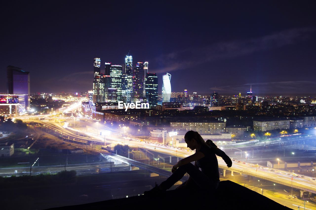 Silhouette woman sitting on terrace against illuminated cityscape at night