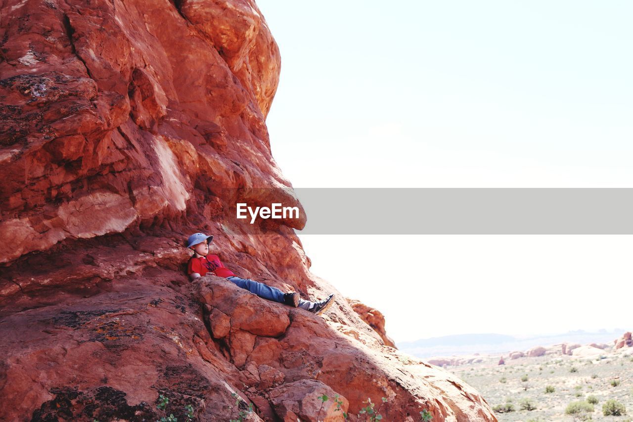 Low angle view of boy relaxing on cliff against clear sky