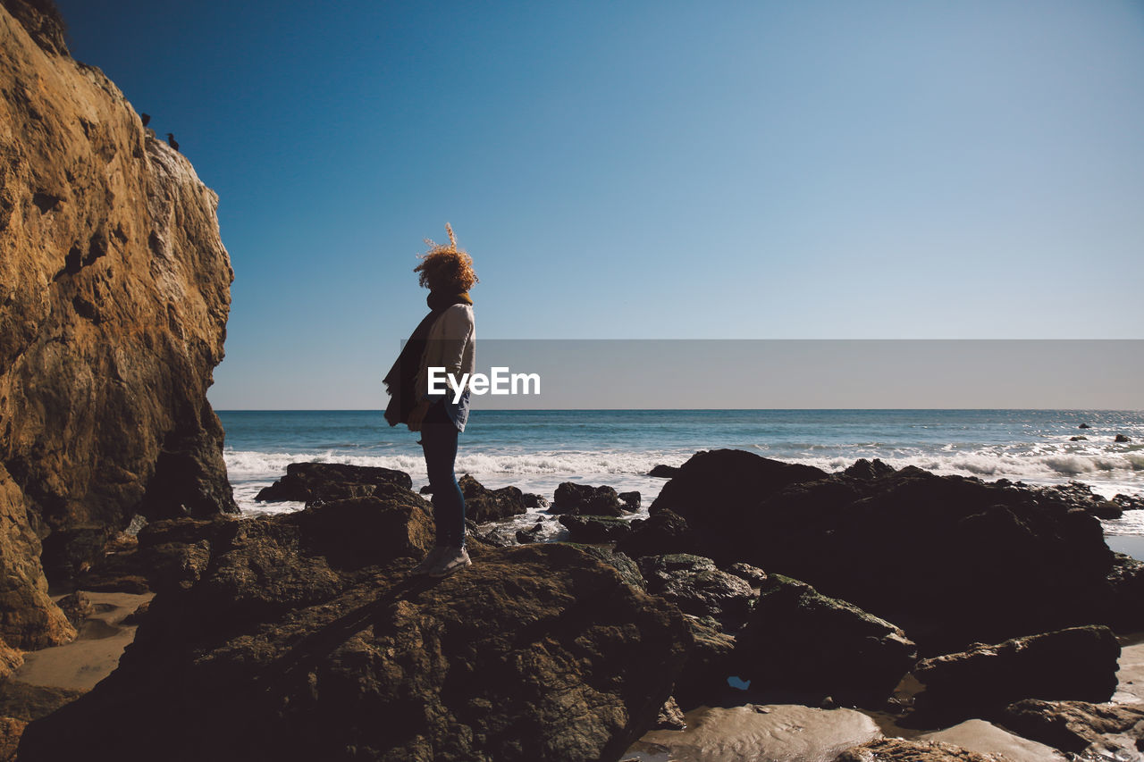 Side view of mid adult woman standing on rocky shore against sky