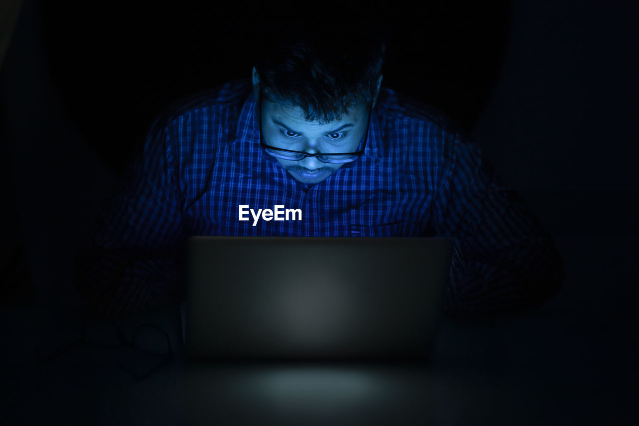 Young man typing on laptop keyboard at night and looking over the eyeglass