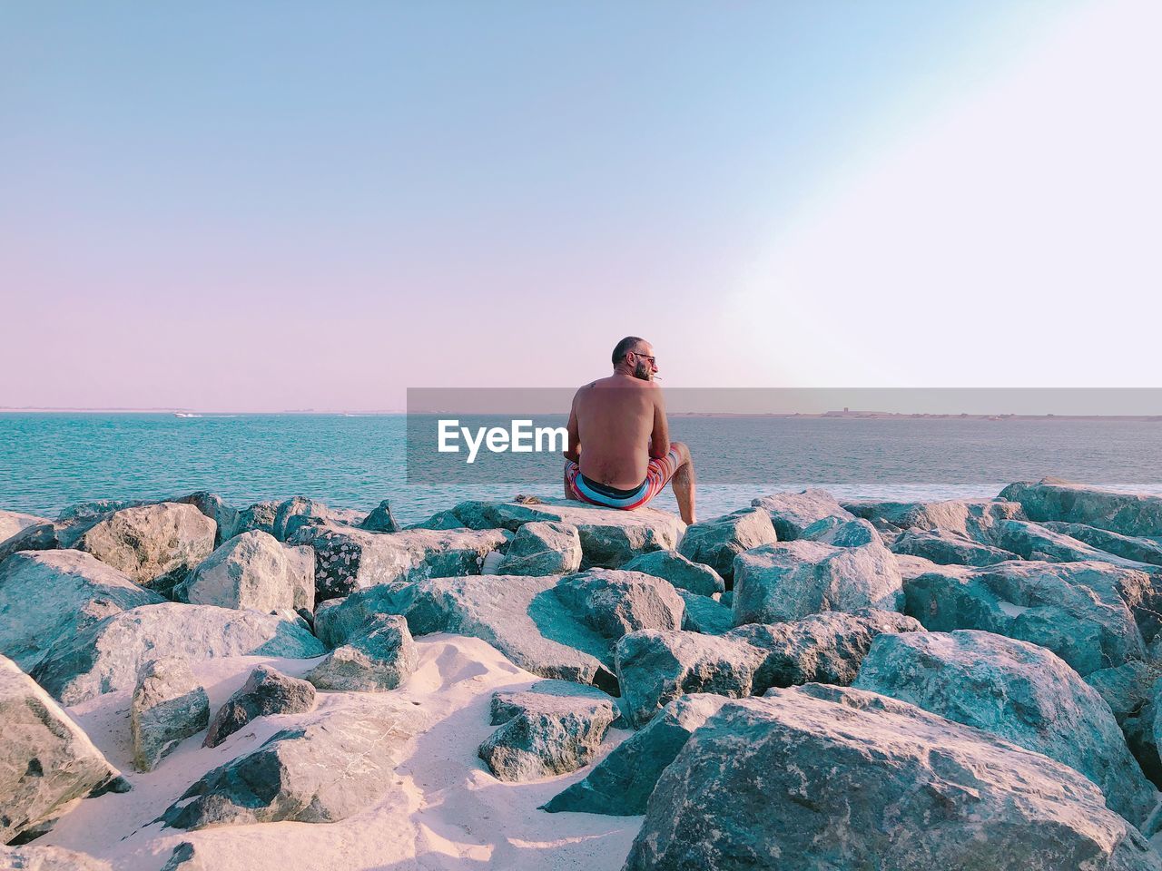 Rear view of shirtless man sitting on rocks by sea against sky