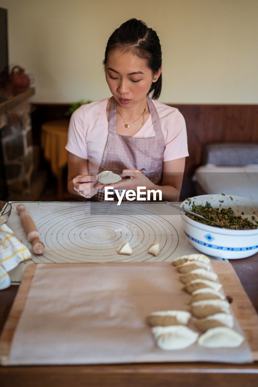 Woman in casual clothes and apron stuffing dumplings with meat while preparing traditional chinese jiaozi in kitchen