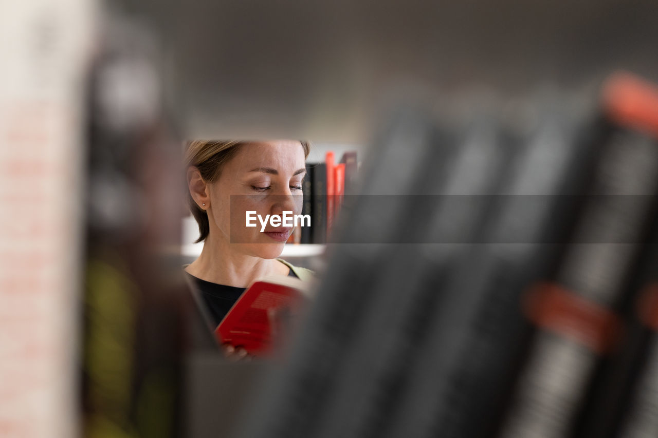 Focused female mature student picking book from bookshelf for educational project preparing for exam