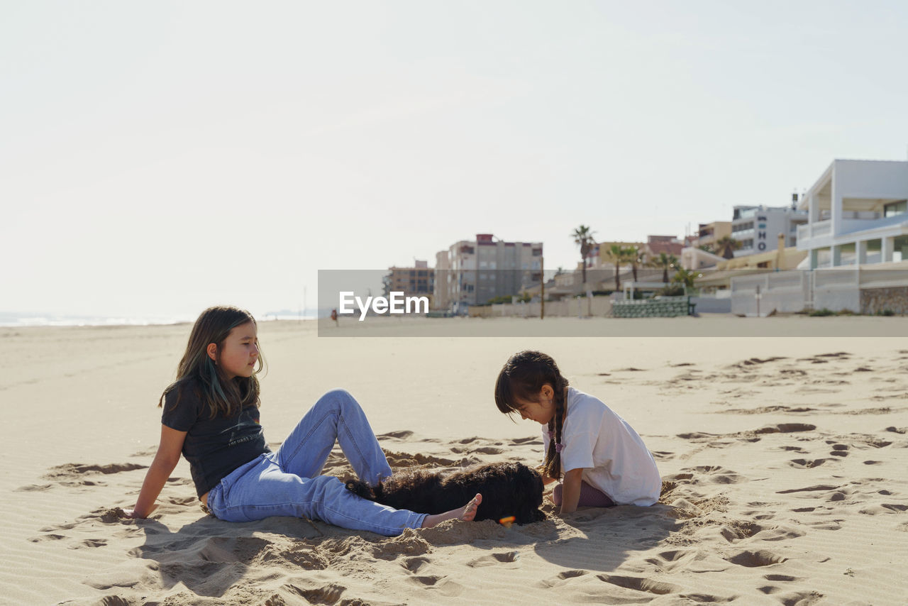 Side view of two girls sitting on beach and playing with dog