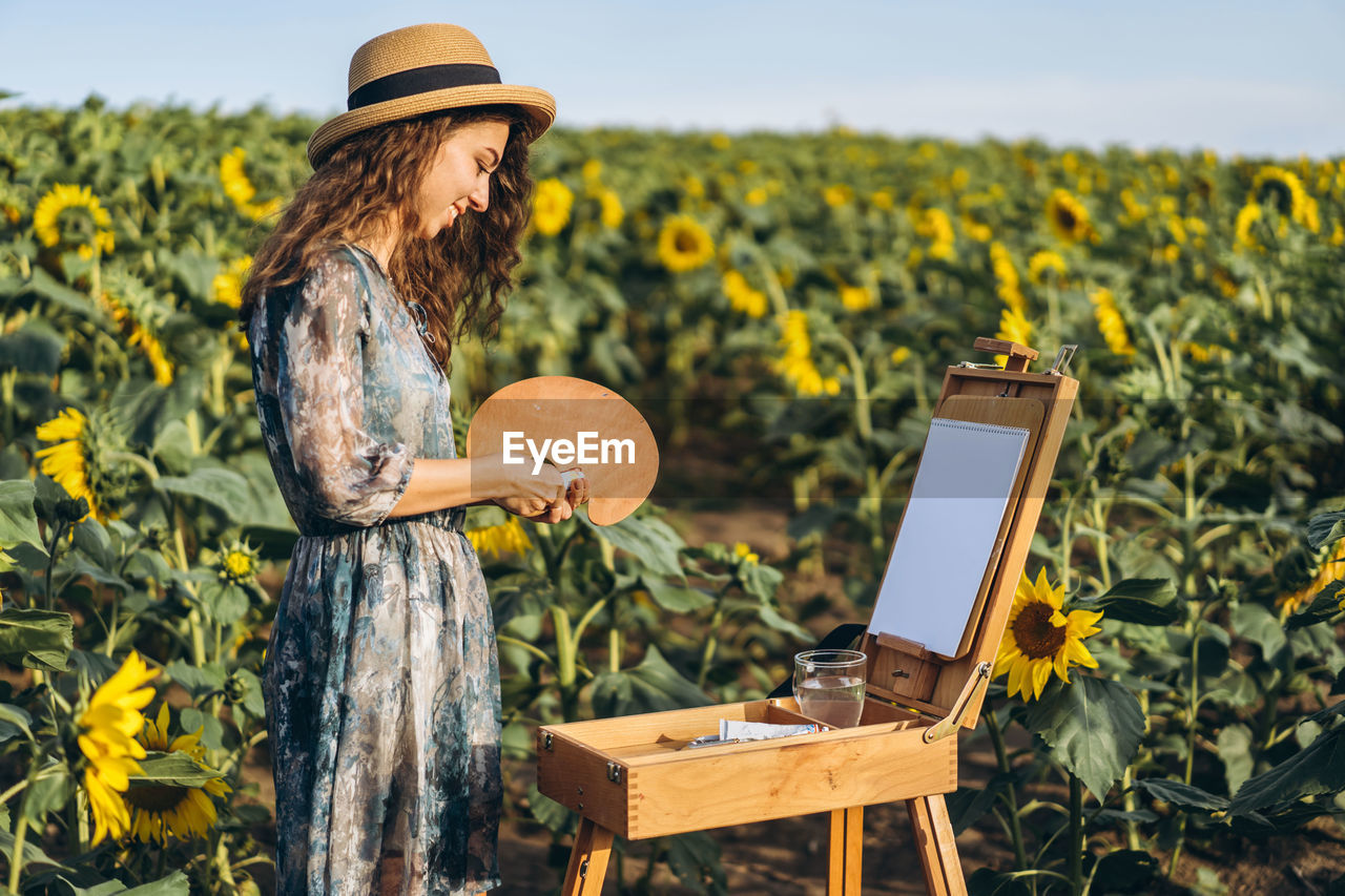 A young woman with curly hair and wearing a hat is painting in nature. 