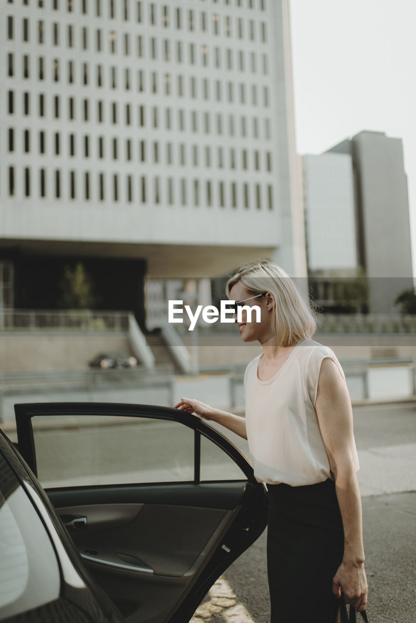 Smiling woman holding car door while standing against building in city