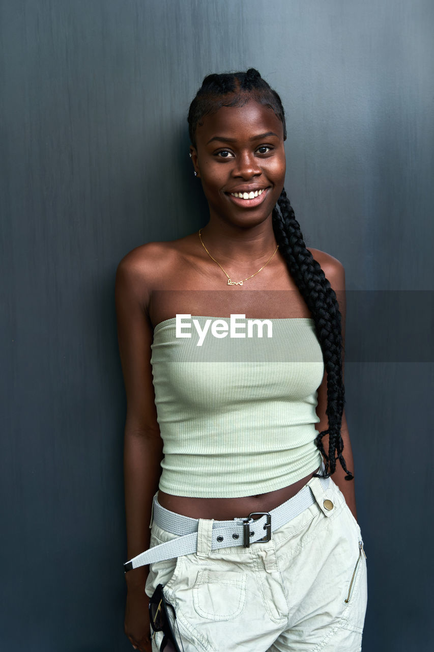 Positive young african american female with long braids wearing tube top with cargo pants and looking at camera on gray background