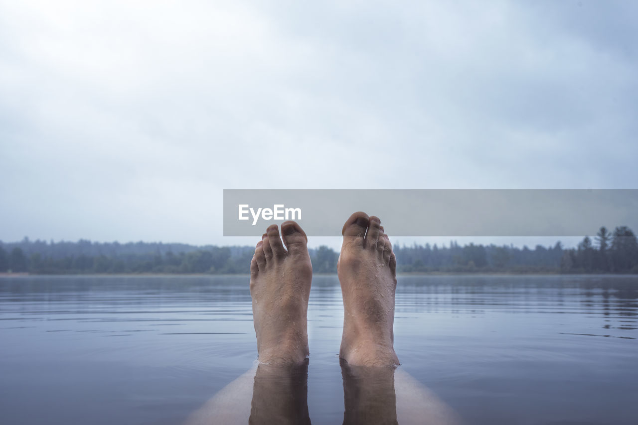 Low section of person relaxing in lake against cloudy sky