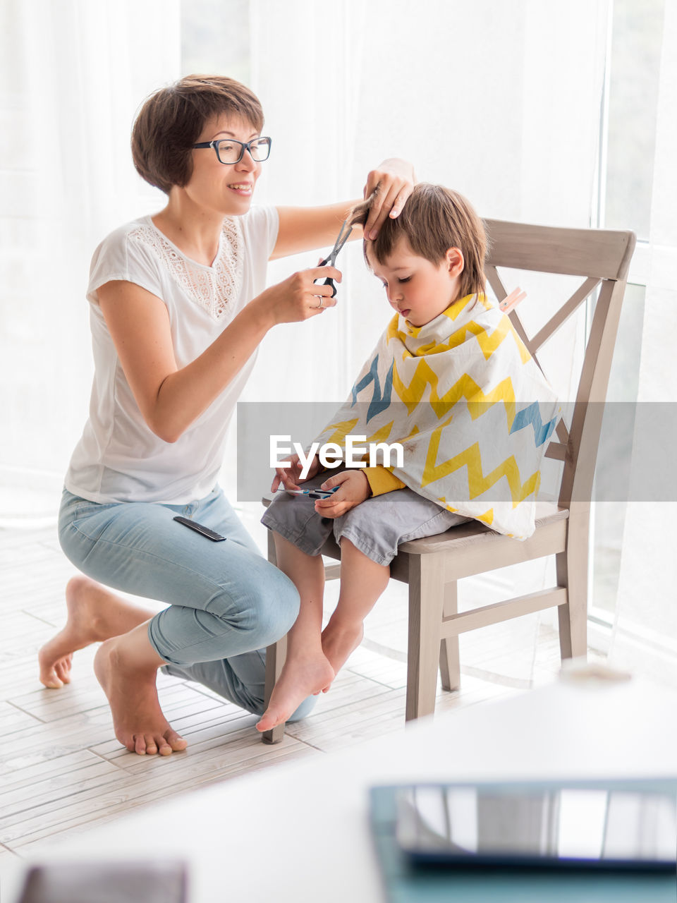 Mother cuts her son's hair by herself. little boy sits, and holds pair of scissors. family life.