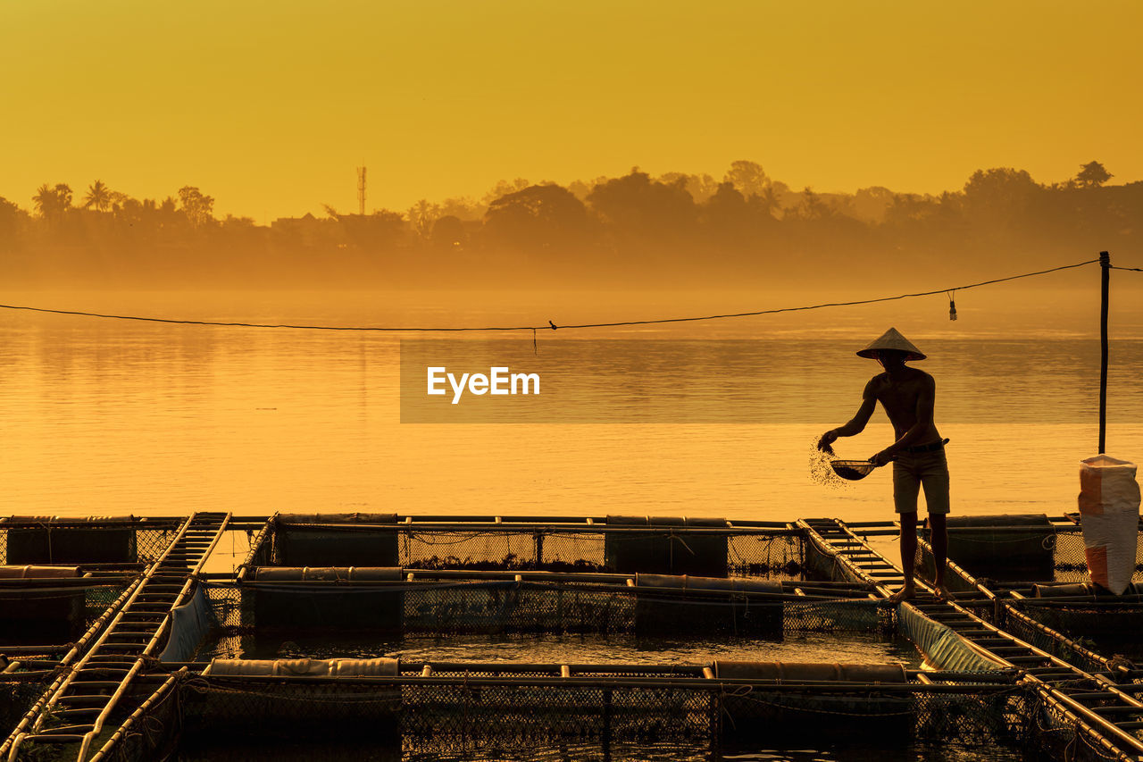 MAN FISHING IN LAKE AGAINST SKY DURING SUNSET