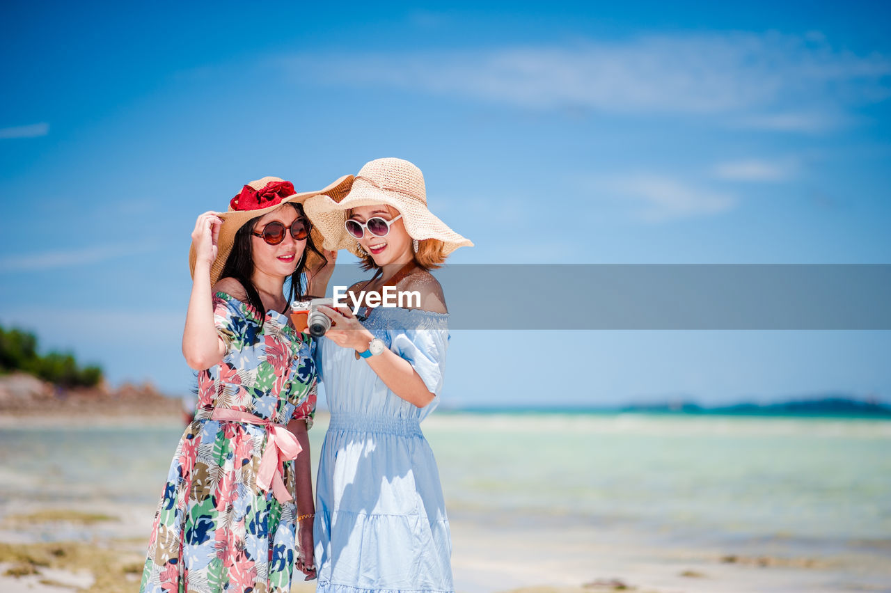 Smiling women using camera while standing at beach against sky