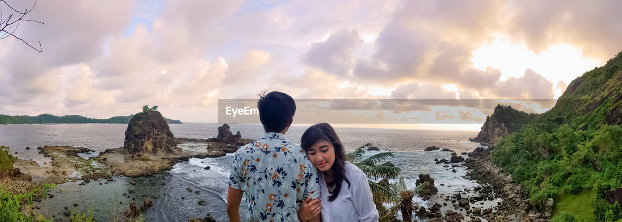 Young couple standing at beach against sky during sunset