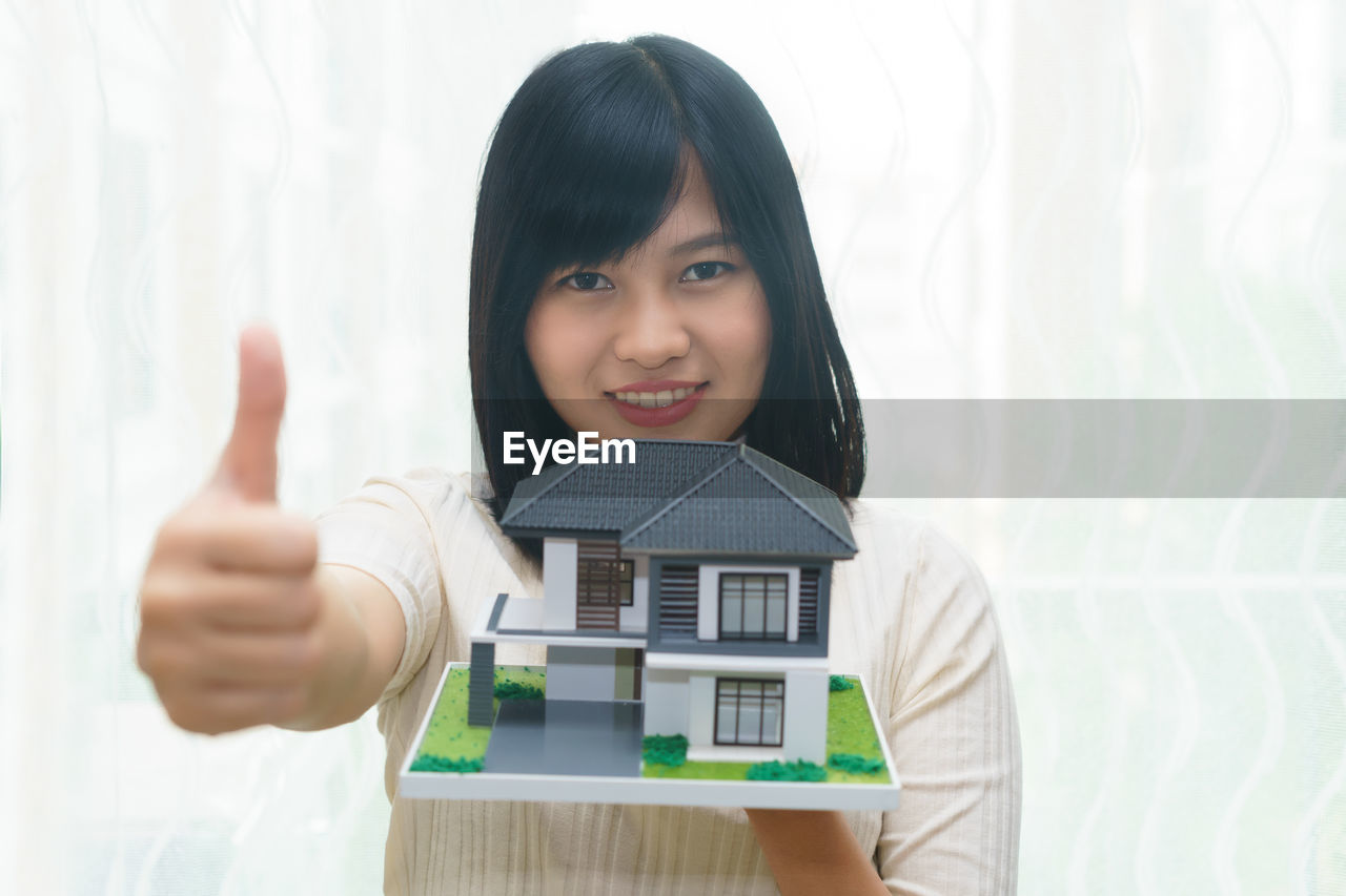 Portrait of smiling young woman holding model house while showing thumbs up