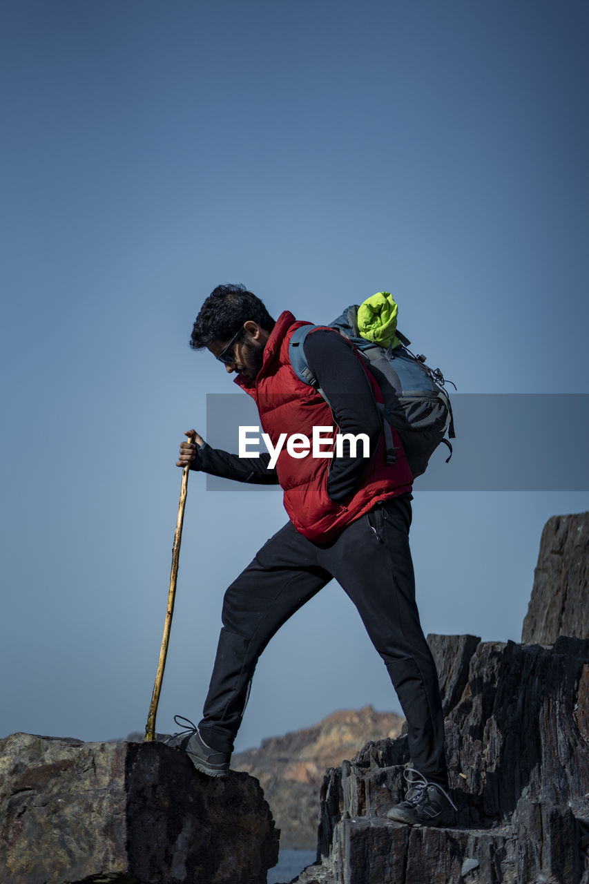 Young indian traveler hiking up the mountain with a backpack and a stick, with a beautiful lake