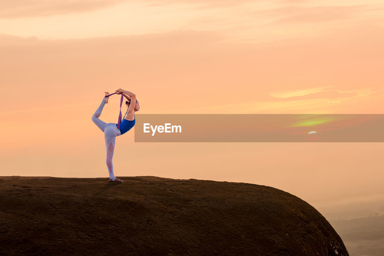 Side view of woman practicing yoga on cliff against orange sky