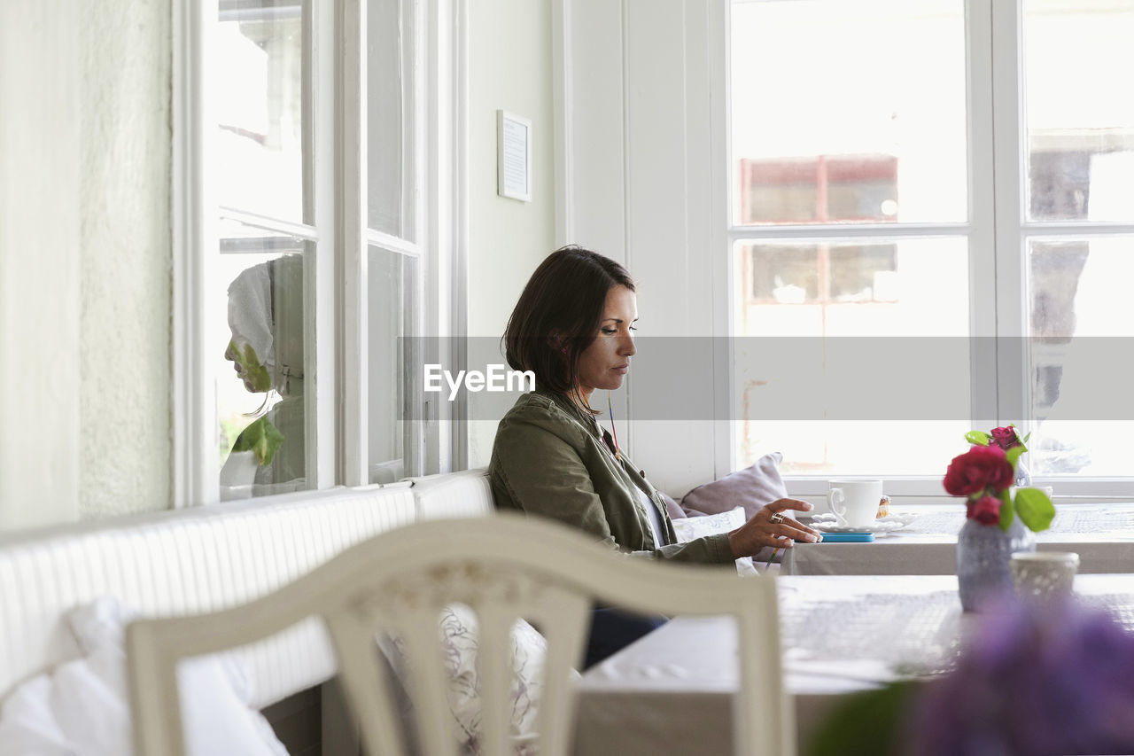 Side view of woman using phone and having coffee while resting at cafe