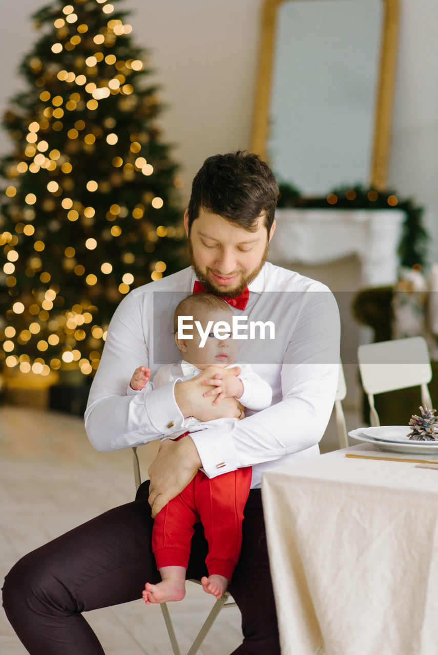 A stylish dad in a white shirt with a red bow tie holds his son in his arms against the background