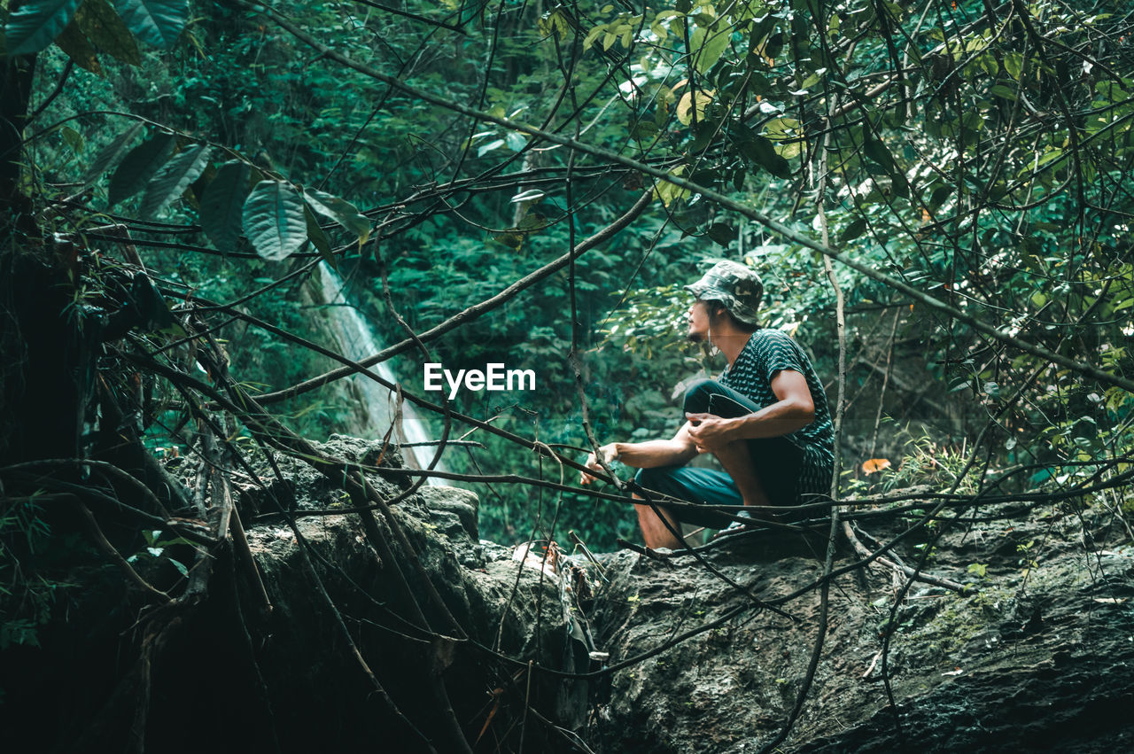 Low angle view of young man sitting on rock in forest