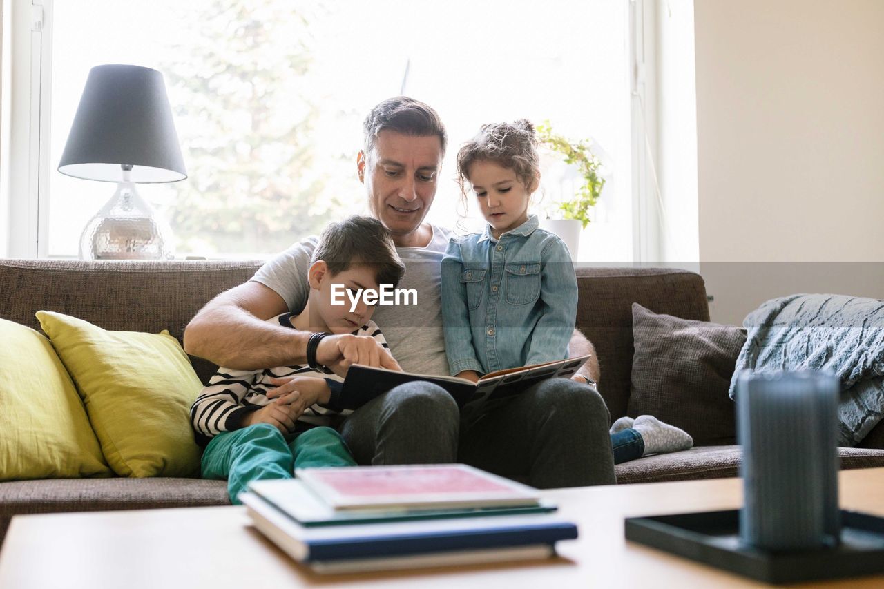 Father reading book to son and daughter while sitting on sofa at home